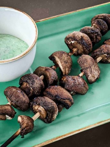 Photo of two skewers of grilled cremini mushrooms with a small bowl of yogurt-herb dressing