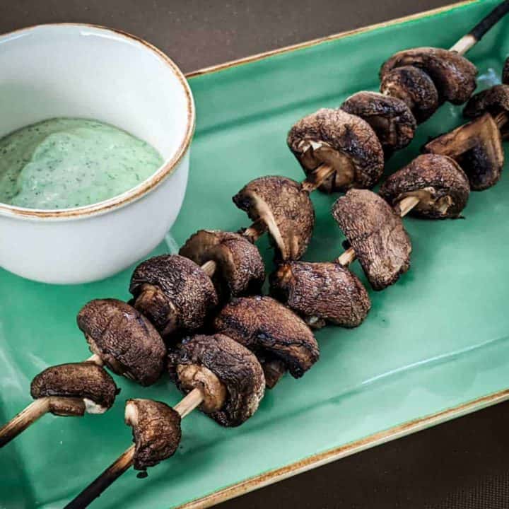 Photo of two skewers of grilled cremini mushrooms with a small bowl of yogurt-herb dressing
