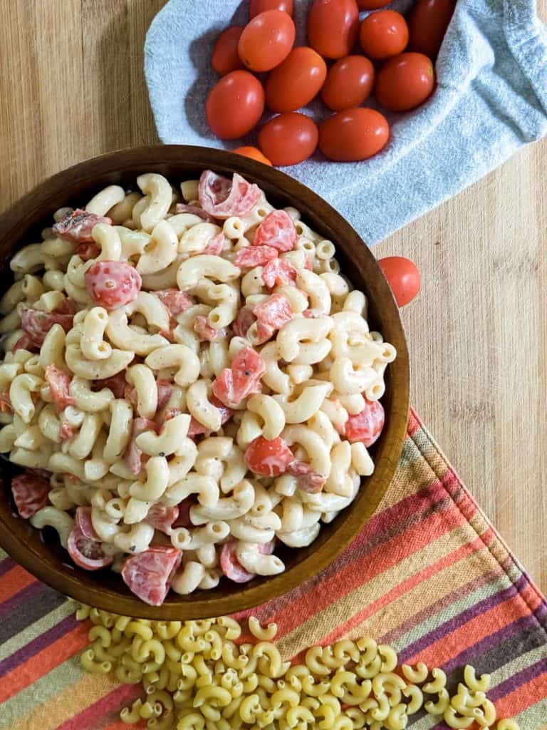 Overhead photo of a large wooden bowl full of macaroni salad on a colorful place mat with fresh cherry tomatoes and dry pasta on the table