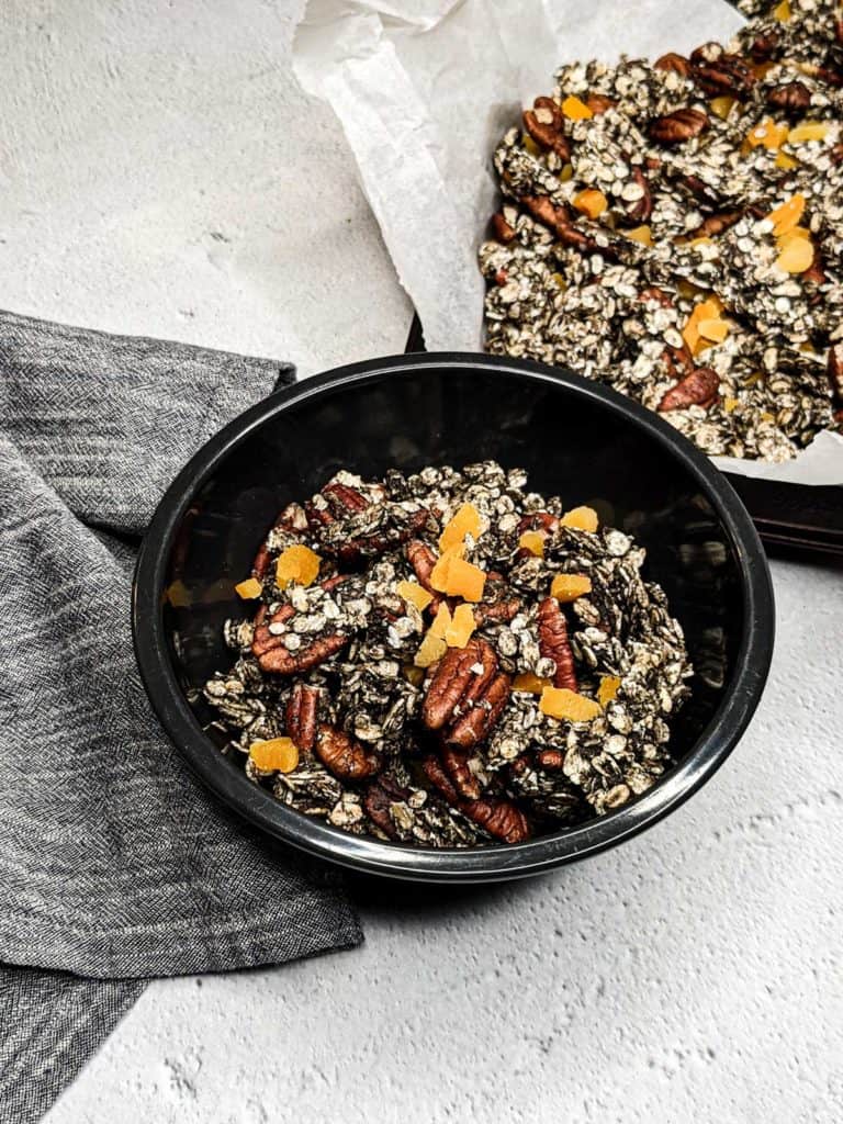 Overhead photo of a bowl of black sesame granola next to a sheet pan lined with parchment paper, filled with the same granola.