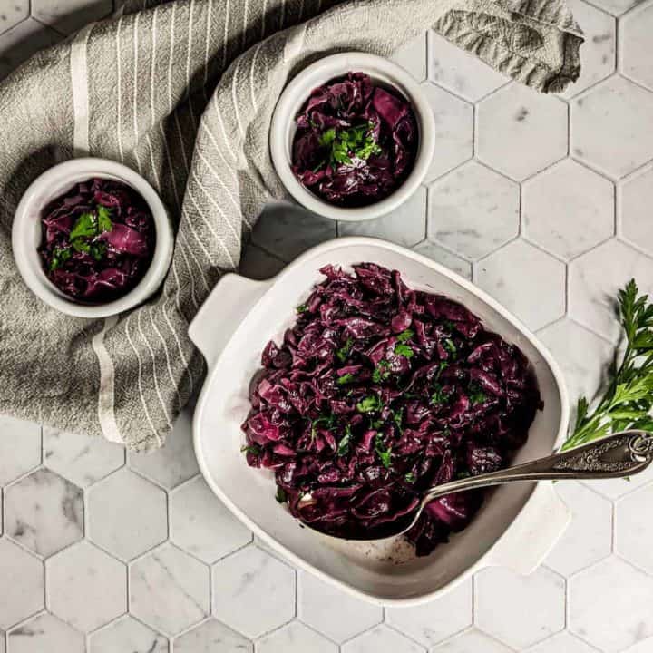 Overhead photo of a serving dish and two individual portions of braised red cabbage with caraway seeds