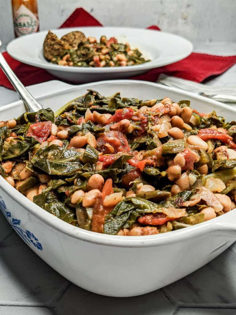 Photo of a serving dish of black eyed peas and collard greens next to a plate with a serving along with rice and cornbread