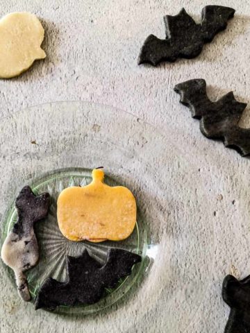 overhead photo of a plate holding a marzipan pumpkin, cat, and bat, next to marzipan bats, cat, and ghost on the table nearby