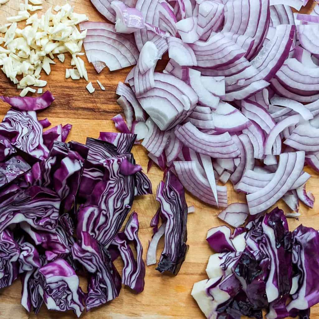 overhead photo of minced garlic, sliced onions and red cabbage on a wooden cutting board