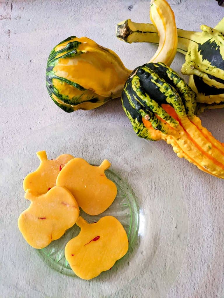 overhead photo of a plate with four saffron-rosewater marzipan pumpkins next to a group of assorted colorful gourds