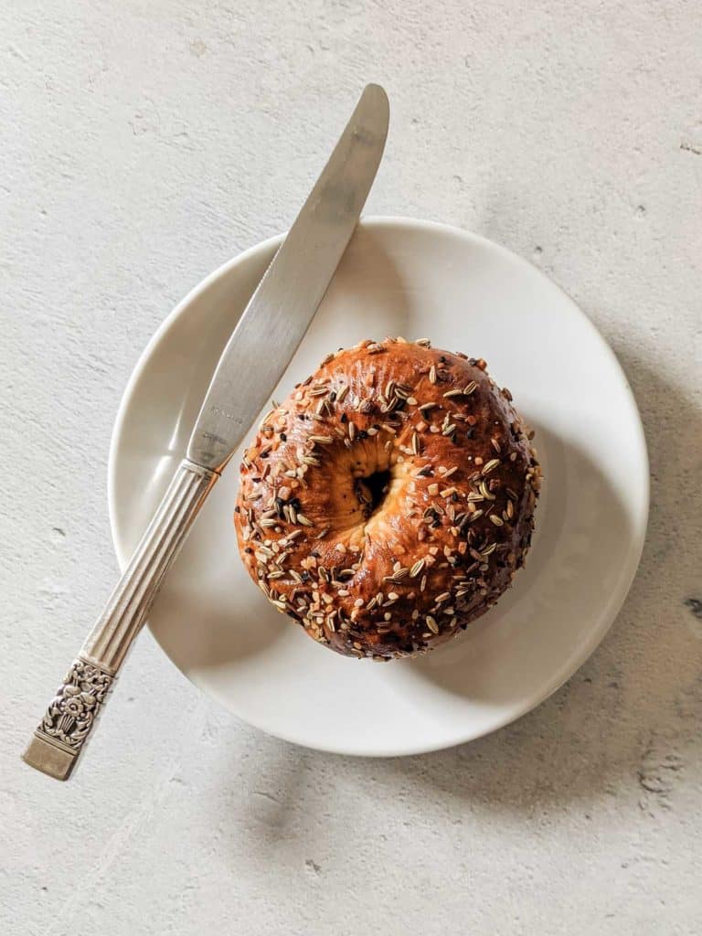 Overhead photo of a bagel on a white plate with a knife